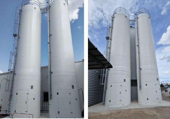 Instalación de dos silos industriales rígidos blancos uno al lado del otro, equipados con escaleras, pasarela y tuberías, delante de un edificio bajo un cielo azul.