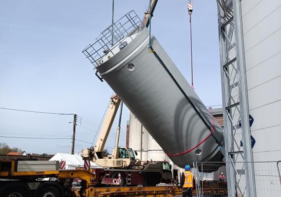 Operario con chaleco de alta visibilidad guiando la instalación de un silo industrial rígido inclinado mediante una grúa móvil montada sobre un camión plataforma en una planta de producción.