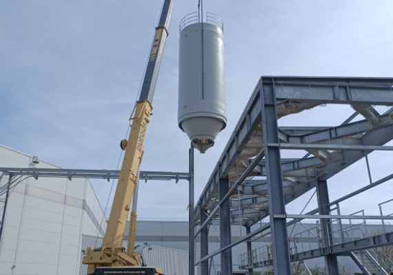 Grúa móvil colocando un silo industrial rígido blanco sobre una estructura metálica exterior en una planta de producción.