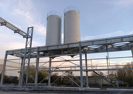Dos silos industriales rígidos blancos instalados sobre una estructura metálica exterior con pasarela y estructura de acceso, fotografiados al atardecer en una planta de producción.
