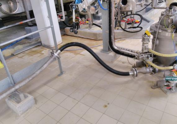 Conexión de tubos de transferencia neumática flexibles negros y tubos inoxidables sostenidos por una estructura metálica sobre el suelo embaldosado.