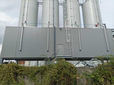 Cuatro silos industriales rígidos blancos alineados sobre una plataforma metálica con escalera de seguridad y tuberías, delante de una fachada con paneles grises.