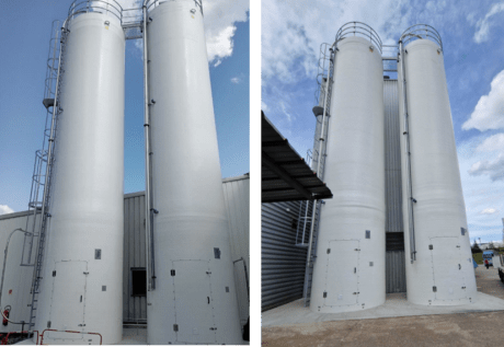 Instalación de dos silos industriales rígidos blancos uno al lado del otro, equipados con escaleras, pasarela y tuberías, delante de un edificio bajo un cielo azul.
