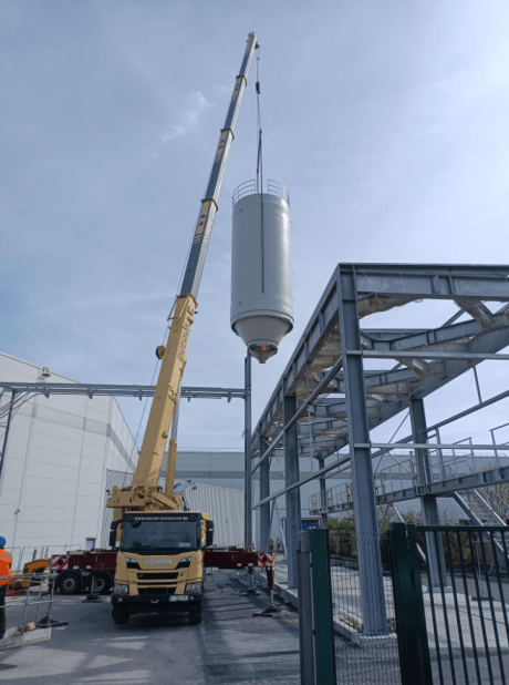 Grúa móvil colocando un silo industrial rígido blanco sobre una estructura metálica exterior en una planta de producción.
