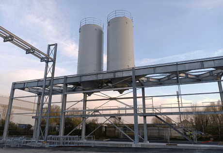 Dos silos industriales rígidos blancos instalados sobre una estructura metálica exterior con pasarela y estructura de acceso, fotografiados al atardecer en una planta de producción.