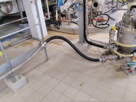 Conexión de tubos de transferencia neumática flexibles negros y tubos inoxidables sostenidos por una estructura metálica sobre el suelo embaldosado.
