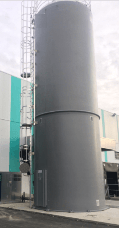 Silo rígido industrial gris antracita equipado con una escalera y una barandilla de acero, instalado delante de una fachada con paneles blancos y turquesa.