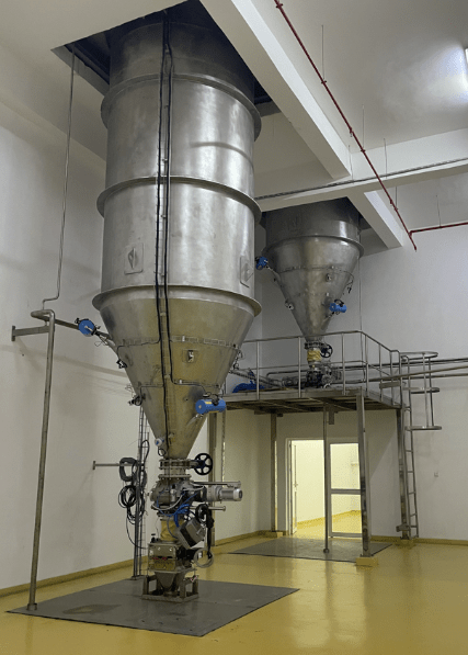 Dos silos rígidos de acero inoxidable instalados en interiores sobre una plataforma metálica con válvulas y tuberías para la alimentación y regulación de un proceso industrial.