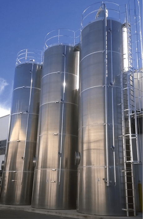 Tres silos industriales rígidos de acero inoxidable brillante, equipados con escaleras y barandillas, instalados en el exterior bajo un cielo azul.
